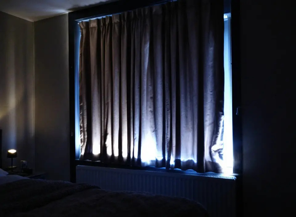 Een slaapkamer in de avond met dichte gevoerde gordijnen voor het raam, een radiator eronder en een brandend nachtlampje